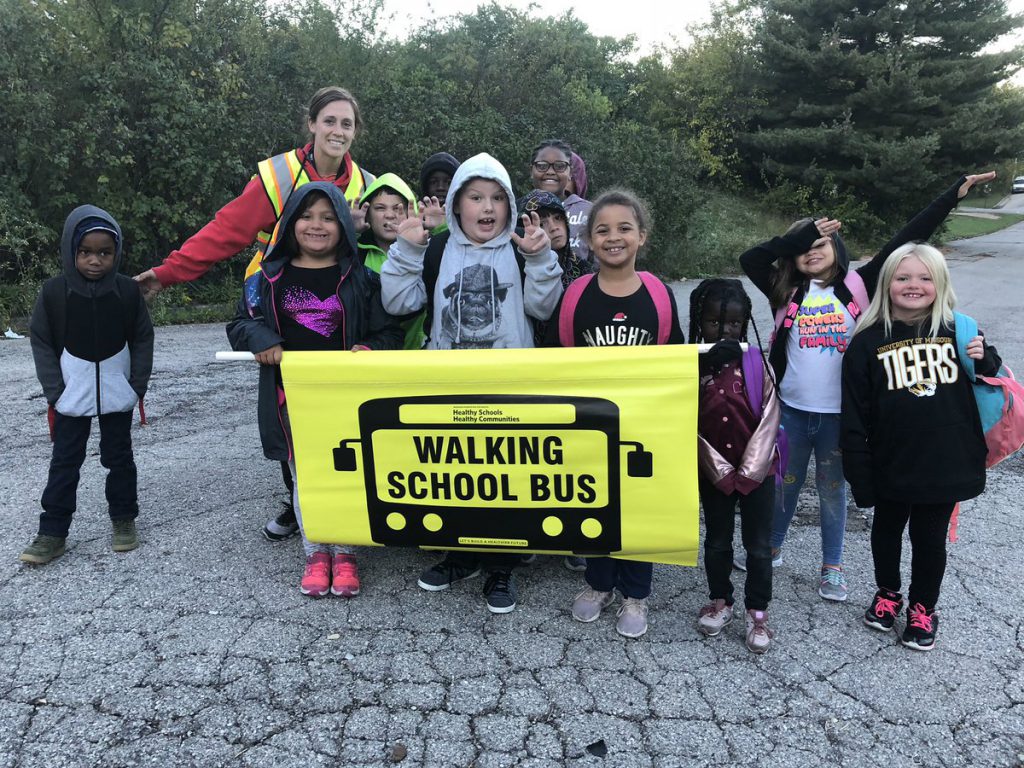 Children and adults stand behind a sign that reads "walking school bus."