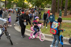 A woman is holding the seat of a child riding a bicycle with training wheels. Around them are other parents at students on the school campus.
