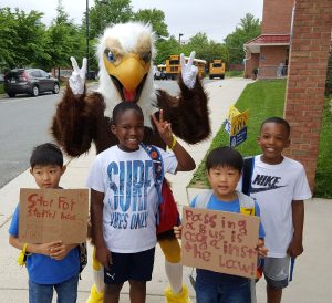 Four children stand in front of a school with an eagle mascot.