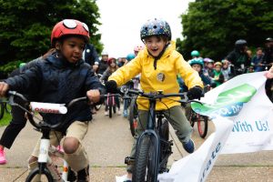 Two children ride bikes through a banner for Bike & Roll to School Day.